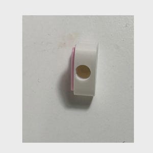 May include: A small, rectangular white plastic component featuring a rounded side and a single circular opening on its face. A thin, light pink accent layer is visible along the left edge of the object, providing a subtle contrast to the white body.