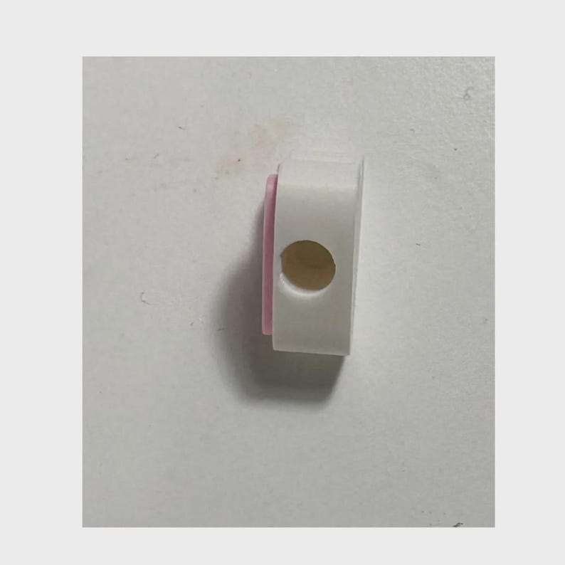 May include: A small, rectangular white plastic component featuring a rounded side and a single circular opening on its face. A thin, light pink accent layer is visible along the left edge of the object, providing a subtle contrast to the white body.