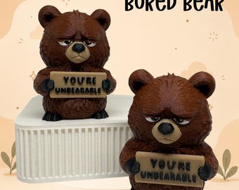 May include: Two brown bear figurines with textured fur, black noses, and grumpy expressions. Each bear holds a tan rectangular sign with the black text 'YOU'RE UNBEARABLE' printed on it. The text 'BORED BEAR' appears in black above the figurines.