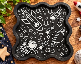 May include: A black square tray with wavy edges featuring a white line art design of a rocket ship, an astronaut, a planet with rings, and various stars and comets scattered across the surface in a space-themed pattern.