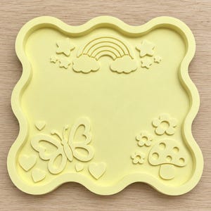 May include: A pale yellow square tray with wavy edges featuring embossed designs on its surface. The tray displays a rainbow with clouds and stars at the top, a butterfly with hearts on the bottom left, and flowers with a mushroom on the bottom right.