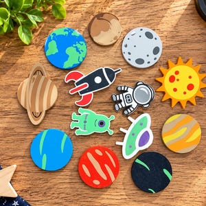May include: A collection of space-themed wooden tokens featuring illustrations of planets, a rocket ship, an astronaut, an alien, and a flying saucer. The set includes a bright yellow sun, a blue Earth, a red Mars, and other celestial bodies.