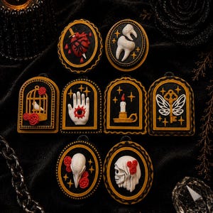 May include: A selection of gothic-style pendants featuring a heart and dagger, a hand with a wound, a bird in a cage, and a skull. Each pendant has a black backdrop with gold edging and a detailed, three-dimensional design, perfect for alternative fashion.