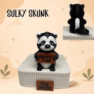May include: A black and white skunk figurine holding a brown sign that reads Stinkin' Tired. A second brown sign on the base reads This Stinks. The text Sulky Skunk appears above. The skunk has a white stripe on its back and a bushy tail.