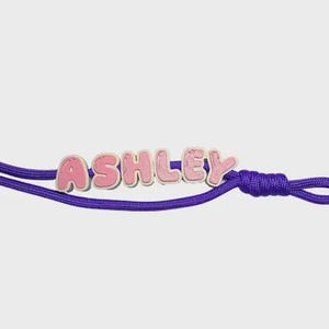 May include: A purple cord bracelet featuring individual light pink letter beads that spell out the name ASHLEY. The letters have a rounded, bubble-like font style and are strung directly onto the vibrant purple cord, which includes a knot detail.
