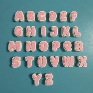 May include: A set of twenty-six individual three-dimensional letters from A to Z, each featuring a white outer border and a pink center with a bubble-style font. A separate white square-shaped object with pink horizontal lines is positioned above.