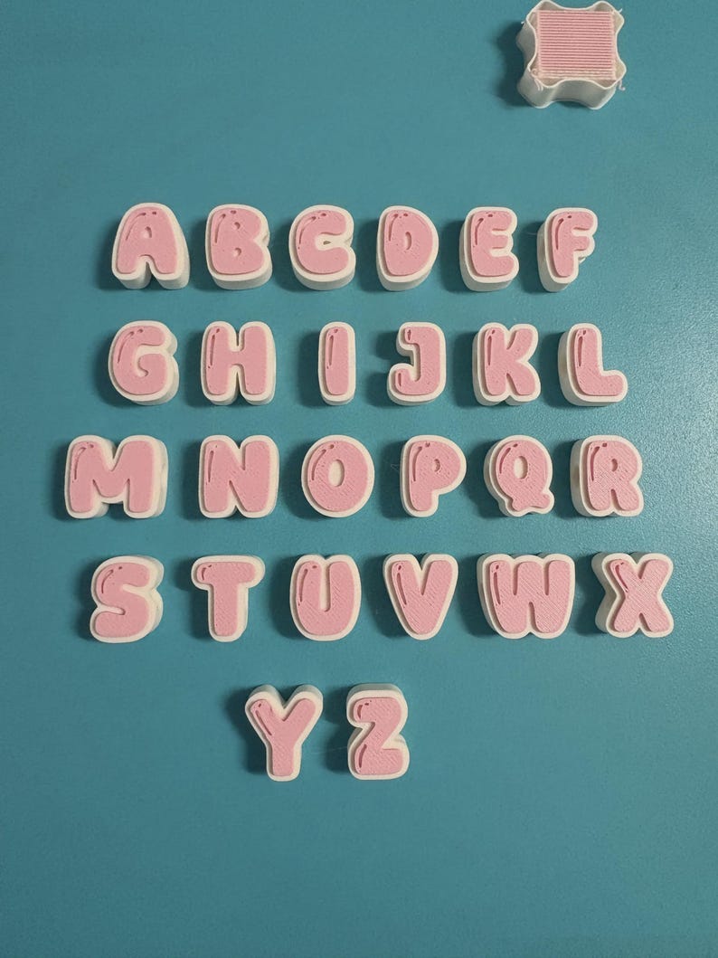 May include: A set of twenty-six individual three-dimensional letters from A to Z, each featuring a white outer border and a pink center with a bubble-style font. A separate white square-shaped object with pink horizontal lines is positioned above.