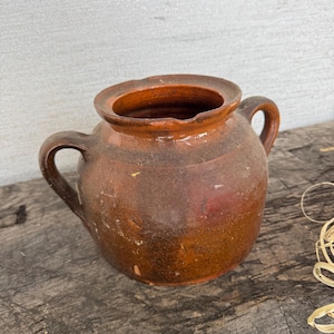 Puede incluir: Una rústica olla de cerámica marrón con dos asas curvas y una abertura ancha. La olla tiene un aspecto ligeramente desgastado con variaciones de color, desde un marrón más claro hasta un marrón rojizo más oscuro. El borde es de un tono más oscuro.
