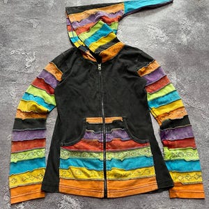 Pode incluir: Um casaco preto com capuz e fecho de correr, capuz pontiagudo e mangas compridas com tiras horizontais de tecido com bordas desfiadas em tons de laranja, amarelo, verde, azul e roxo. A frente inclui um bolso canguru dividido com tiras coloridas.