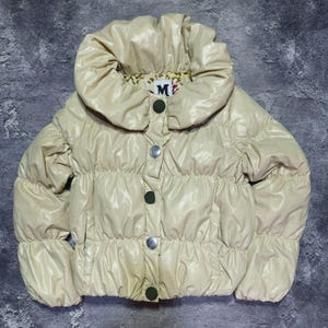 May include: A cream-colored puffer jacket with a large, rounded collar. The jacket features a button-up front with silver and dark green buttons. The brand logo "M" is visible on the collar lining. The jacket has a quilted design.