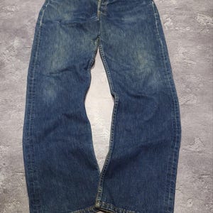 May include: A pair of dark blue denim jeans featuring a button-fly closure, five-pocket styling, and a straight-leg cut. The fabric displays a subtle faded wash pattern throughout the front panels, with visible stitching along the seams and hems.