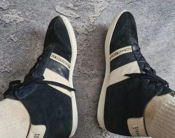 Pode incluir: Um par de ténis de cano alto azul marinho com detalhes e atacadores brancos. Os ténis apresentam uma faixa branca na lateral e o nome da marca "Bikkembergs" em branco. As solas são brancas.