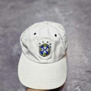 May include: White baseball cap featuring the Brazilian Football Confederation (CBF) emblem. The cap has a curved brim and a structured crown with two small black eyelets. The CBF logo is embroidered in green, blue, and yellow.
