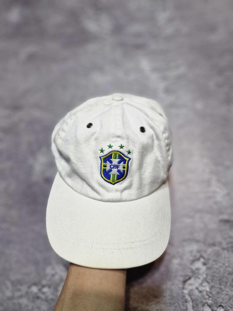 May include: White baseball cap featuring the Brazilian Football Confederation (CBF) emblem. The cap has a curved brim and a structured crown with two small black eyelets. The CBF logo is embroidered in green, blue, and yellow.