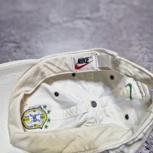 May include: Off-white Nike baseball cap with a curved brim. The interior features a Nike tag with a red swoosh logo. The cap has a Brazil emblem and green embroidered details. The cap is made of a durable fabric.
