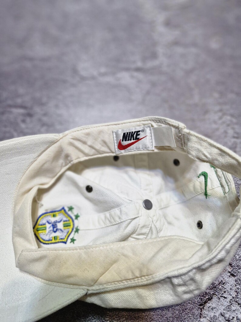 May include: Off-white Nike baseball cap with a curved brim. The interior features a Nike tag with a red swoosh logo. The cap has a Brazil emblem and green embroidered details. The cap is made of a durable fabric.