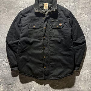 Può includere: Giacca da lavoro nera con chiusura a bottoni, due tasche sul petto e due tasche inferiori. La giacca ha un colletto in velluto a coste e una fodera trapuntata. Il logo Dickies è visibile.