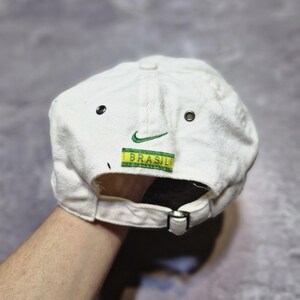 May include: White baseball cap with a green Nike logo and the word "BRASIL" in green and yellow. The cap has a metal buckle closure and two small eyelets on the side.