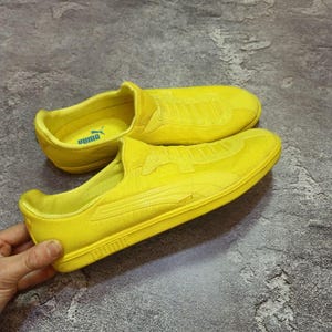 Puede incluir: Zapatillas slip-on de color amarillo brillante. Los zapatos tienen una parte superior de tela texturizada y una suela de goma. El logotipo de Puma es visible en el lateral y en la plantilla. Los zapatos son de un color sólido y vibrante.