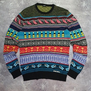 May include: A Diesel sweater with a vibrant, patterned design. The sweater features repeating motifs of citrus slices, bottles, palm trees, and flags in a variety of colors including blue, red, and green. The word "DIESEL" is visible near the bottom.