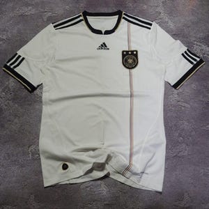 May include: A white Adidas Germany national soccer team jersey featuring black three-stripe detailing on the shoulders, a black crew neckline, the Adidas logo, and the German national team crest with three stars on the chest, plus a vertical pinstripe.