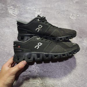 May include: A pair of black athletic sneakers featuring a mesh upper, black laces, and a white On logo on the side. The shoes have a textured black sole with distinct hollow pods, a grey interior lining, and a small Swiss flag detail on the heel.
