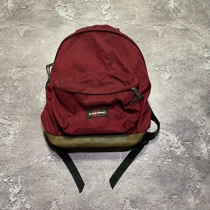 May include: A burgundy backpack featuring a large main compartment and a smaller front zippered pocket. The bag has a dark olive green textured base and black straps. A rectangular black patch with the white text EASTPAK is centered.