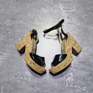 Puede incluir: Un par de sandalias de plataforma negras y de corcho. Las sandalias tienen un diseño de punta abierta con una parte superior de charol negro y un tacón de plataforma de corcho. Las correas del tobillo son negras con una hebilla dorada. Los tacones miden unos 10 cm.