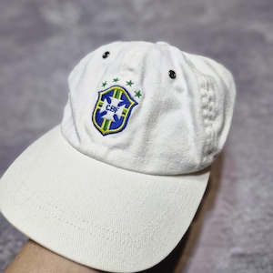 May include: White baseball cap featuring the Brazilian Football Confederation (CBF) logo in blue, green, and yellow. The cap has a curved brim and adjustable strap. The CBF logo is on the front of the cap.