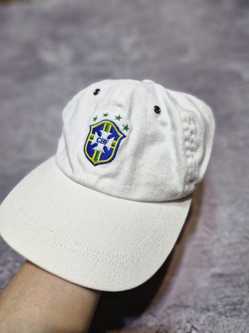 May include: White baseball cap featuring the Brazilian Football Confederation (CBF) logo in blue, green, and yellow. The cap has a curved brim and adjustable strap. The CBF logo is on the front of the cap.