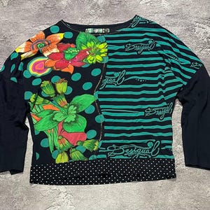 May include: A long-sleeve shirt featuring a split design with a black background, large colorful floral and polka dot patterns on the left, and horizontal green and black stripes with the text Desigual on the right, finished with a white polka dot hem.