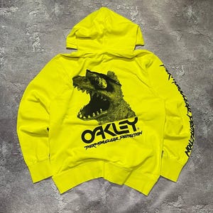 Puede incluir: Una sudadera con capucha de color amarillo neón brillante que presenta en la espalda un gráfico negro de un dinosaurio con gafas de sol. Debajo del dinosaurio aparece el texto negro Oakley Thermonuclear Protection, impreso también en la manga izquierda.