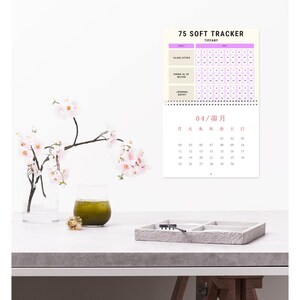 May include: A calendar labelled "75 Soft Tracker" with a light purple and white design, for tracking daily habits. A glass of green liquid, a small tray, and a vase with pink flowers are on a white surface.