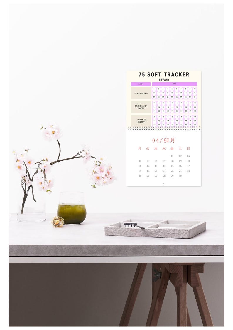 May include: A calendar labelled "75 Soft Tracker" with a light purple and white design, for tracking daily habits. A glass of green liquid, a small tray, and a vase with pink flowers are on a white surface.