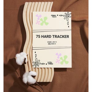 May include: A cream-coloured 75 Hard Tracker card with floral designs and the start and end dates of the challenge. The card is propped up on a wooden, wavy, decorative piece, with cotton stems in the foreground.
