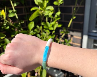 May include: A cylindrical bangle bracelet featuring three distinct colour segments in a smooth, glossy finish. The bracelet is composed of a solid white section, a vibrant bright blue section, and a translucent sage green section joined together.