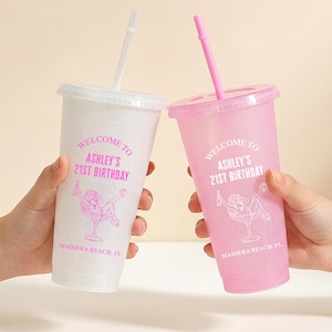 May include: Two glittery plastic tumblers with lids and straws, one white and one pink. Each cup features pink text reading "WELCOME TO ASHLEY'S 21ST BIRTHDAY" and "MADIERA BEACH, FL" above a graphic of a person lounging in a large cocktail glass.