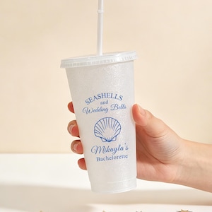 May include: A clear, glitter-covered plastic cup with a lid and straw. The cup features blue text that reads "Seashells and Wedding Bells" and "Mikayla's Bachelorette" with a seashell graphic. The cup is held by a person.