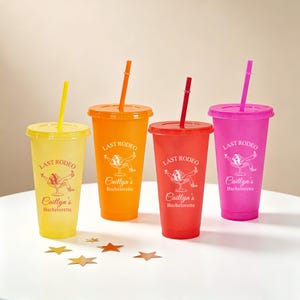 K&ouml;nnte beinhalten: Vier bunte Plastikbecher mit Deckeln und Strohhalmen. Die Becher sind gelb, orange, rot und pink. Jeder Becher tr&auml;gt den Text "LAST RODEO" und "Caitlyn's Bachelorette". Goldfarbene sternf&ouml;rmige Konfetti sind auf dem Tisch verstreut.