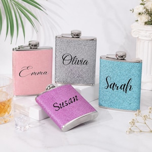 Peut inclure: Quatre flasques en acier inoxydable avec des finitions pailletées roses, argentées, violettes et bleues. Chaque flasque porte un nom en cursive : Emma, Olivia, Susan et Sarah. Les flasques ont un bouchon argenté et une forme rectangulaire.