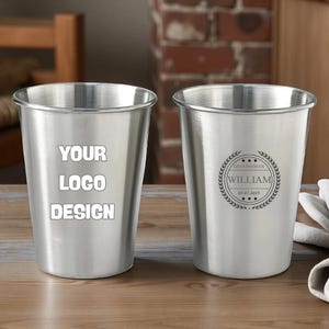 Puede incluir: Dos vasos de acero inoxidable. Uno presenta el texto "YOUR LOGO DESIGN" en blanco. El otro tiene un diseño circular con la palabra "WILLIAM" y la fecha 01.07.2023, rodeado por una corona de laurel.