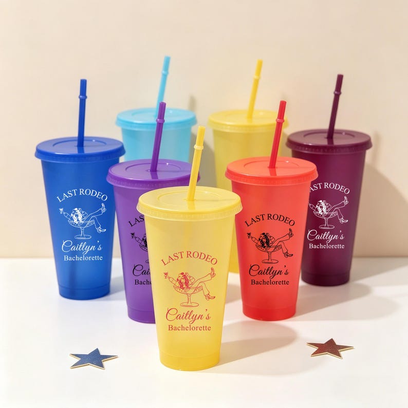 K&ouml;nnte beinhalten: Eine Sammlung bunter Plastikbecher mit Deckeln und Strohhalmen. Jeder Becher hat eine andere Farbe, darunter Blau, Lila, Gelb, Rot und Burgunder. Der Text "LAST RODEO Caitlyn's Bachelorette" ist auf jedem Becher aufgedruckt.