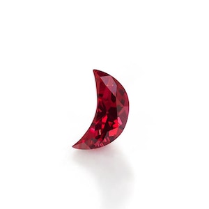 May include: A crescent moon-shaped gemstone with a deep red colour and a faceted surface. The stone features multiple triangular and diamond-shaped facets that reflect light, creating a brilliant appearance against a plain white background.