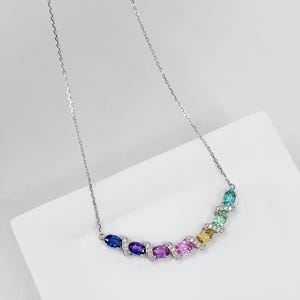 Peut inclure: Un collier argenté doté d'un pendentif incurvé orné de sept pierres précieuses ovales multicolores en bleu, violet, rose, jaune, vert clair et sarcelle, accentué par de petites pierres claires sur les maillons et une fine chaîne.