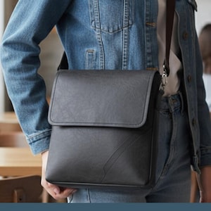 May include: A black messenger bag with a flap closure and a shoulder strap. The bag is made of a smooth material and has a minimalist design. The person is wearing a denim jacket and jeans.