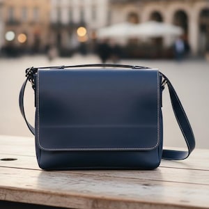 May include: A navy blue messenger bag with a flap closure and a shoulder strap. The bag is made of a smooth material and has white stitching around the edges. It is sitting on a wooden table.