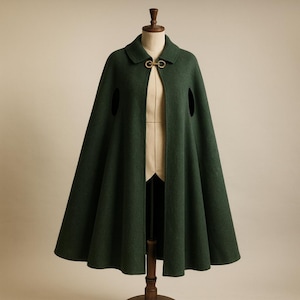 May include: A dark green cape with a collar and a gold-colored clasp. The cape has two arm openings and is displayed on a wooden mannequin. The fabric appears to be a wool blend, and the cape has a flowing design.