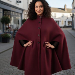 May include: A burgundy cape with a high collar and button closure. The cape features a flared silhouette and arm openings. The woman is wearing a black top underneath. The background shows a brick-paved street and buildings.