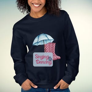 Könnte beinhalten: Marineblaues Sweatshirt mit einem Aquarelldesign eines hellblauen Regenschirms, rosa gepunkteten Gummistiefeln und den Worten "Singing & Dancing" auf hellblauem Hintergrund. Das Sweatshirt ist einfarbig.