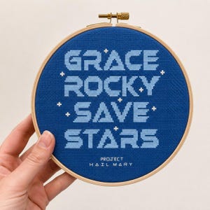 Op de afbeelding: Een ronde houten borduurring met een blauw kruissteek borduurwerk. De tekst GRACE ROCKY SAVE STARS is in lichtblauw geborduurd, geaccentueerd met kleine witte kruisjes. Daaronder staat de tekst PROJECT HAIL MARY in het wit geborduurd.